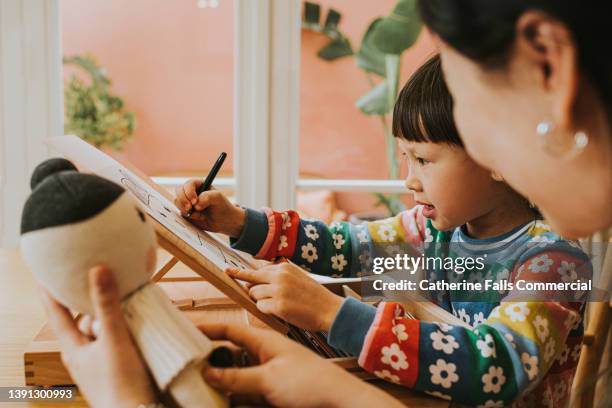 a little girl uses an artists easel on a table to draw, as an adult sits beside her and encourages and engages with the activity. - chinese tutor study stock-fotos und bilder