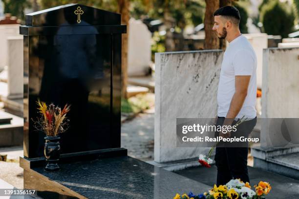 junger mann, der blumen am grabstein auf dem friedhof platziert - begräbnisstätte stock-fotos und bilder