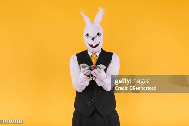 disguised person in rabbit mask with suit jacket, vest and tie, holding easter eggs, on yellow background. carnival, party, easter and celebration concept. - easter bunny mask stockfoto's en -beelden