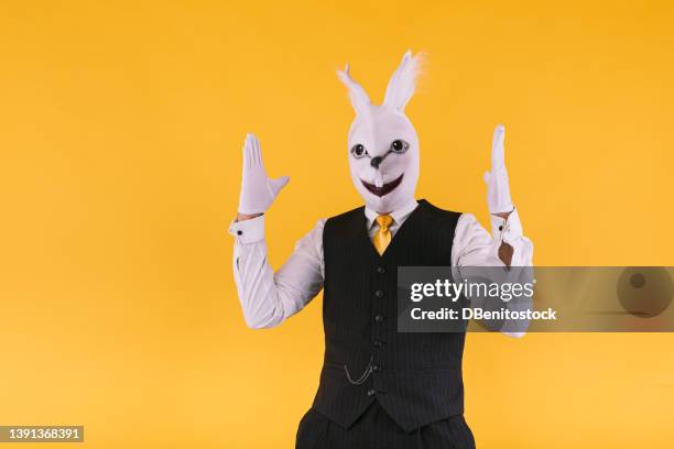 disguised person in rabbit mask wearing suit jacket, vest and tie, raising arms excited, on yellow background. carnival, party, easter and celebration concept. - easter mask stock-fotos und bilder