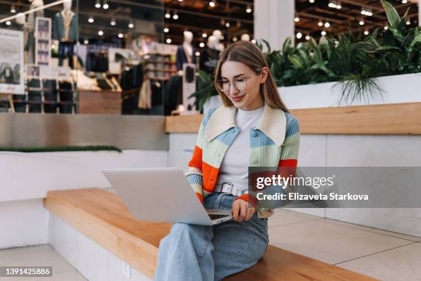 a young female freelancer influencer in glasses and fashionable casual clothes works online remotely using a laptop while sitting on the stairs in a shopping mall in a public place. a girl university student in a bright sweater to study online using - generation z laptop stock pictures, royalty-free photos & images