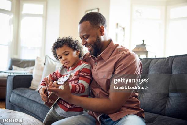 father and son play a ukulele on the sofa. - vater sohn musik stock-fotos und bilder