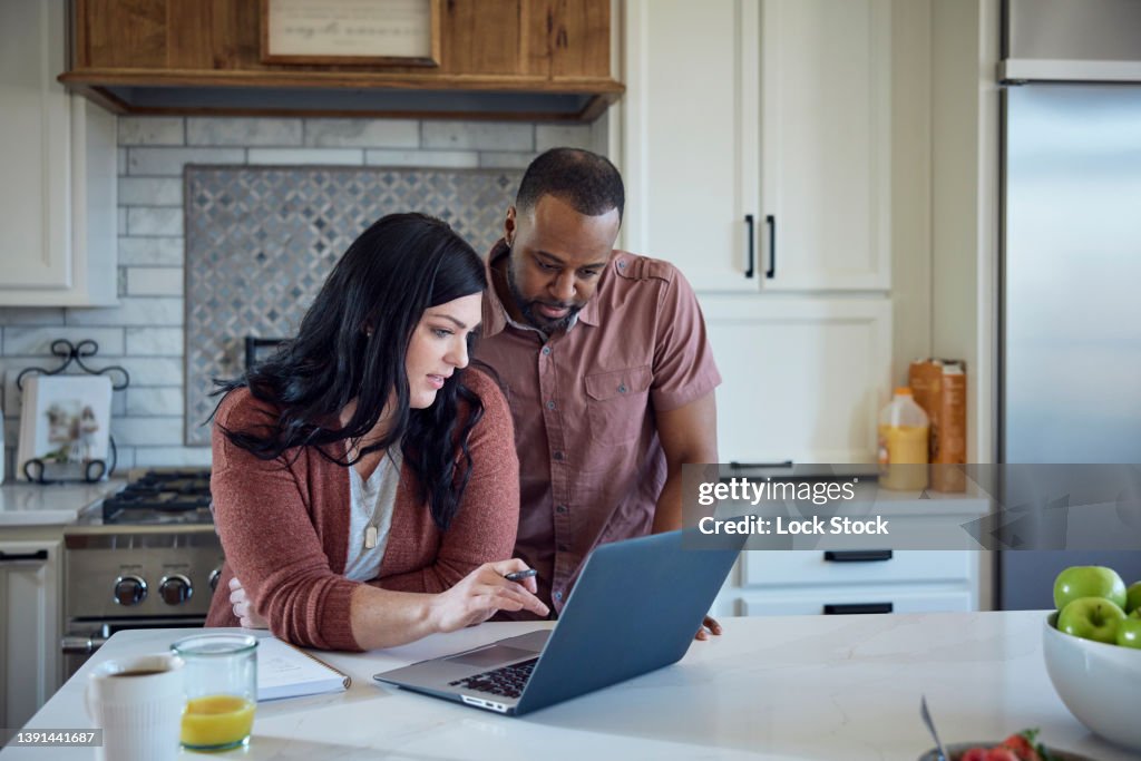 Caucasian woman and African American man sit at kitchen counter with breakfast working with pen, paper and laptop.