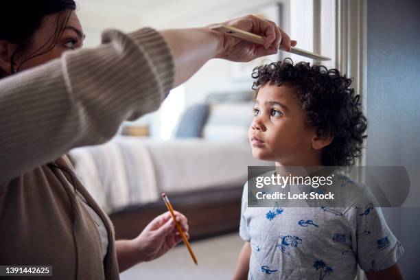 caucasian mother measuring height of mixed-race son on wall. - height chart stock pictures, royalty-free photos & images