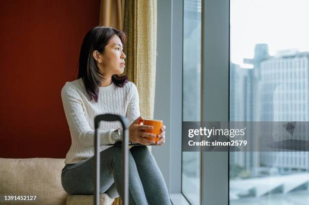 portrait of an asian tourist woman sitting at sofa by the window, holding coffee in hotel room after check-in - solo travel concept - coffee cup top view stockfoto's en -beelden