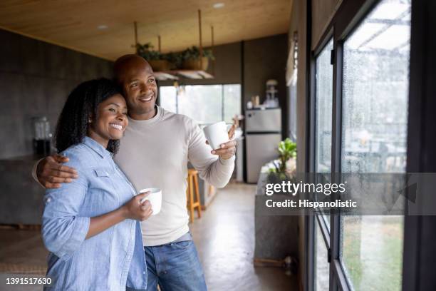 couple at home looking out the window while drinking a cup of coffee - rainy day window stock pictures, royalty-free photos & images