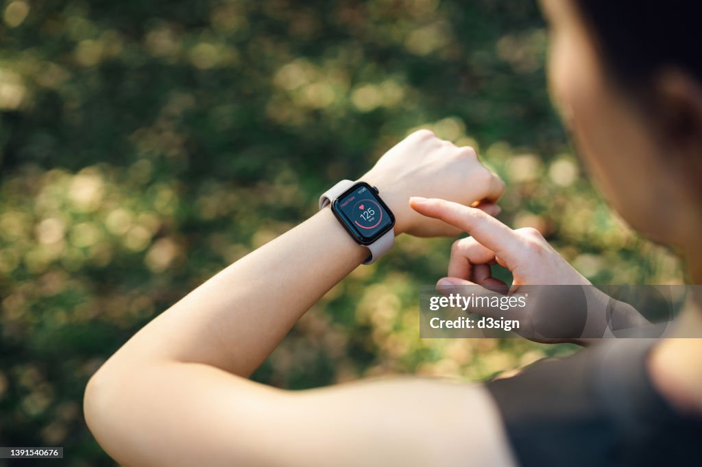 Over the shoulder view of young Asian sports woman resting after working out outdoors in green park, measuring heart rate on her smartwatch. Health and fitness training with technology. Wearable technology. Healthy living lifestyle, sports routine concept