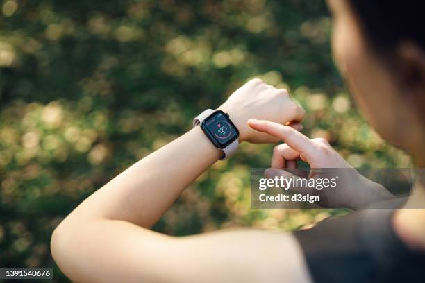 over the shoulder view of young asian sports woman resting after working out outdoors in green park, measuring heart rate on her smartwatch. health and fitness training with technology. wearable technology. healthy living lifestyle, sports routine concept - montre connectée photos et images de collection