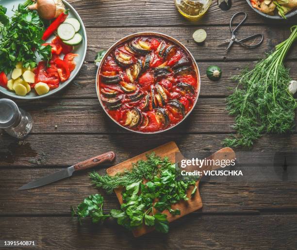 homemade ratatouille with courgette, bell pepper and potato in casserole on rustic wooden kitchen table with cutting board, ingredients and kitchen utensils. - ratatuia foto e immagini stock