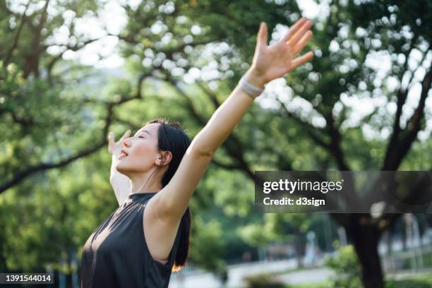 young asian sports woman with eyes closed stretching with arms outstretched after working out in green park outdoors with sunlight in morning. health and fitness training, healthy living lifestyle, sports routine concept - woman home run stock pictures, royalty-free photos & images