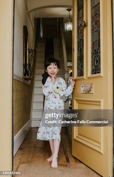 a cute little girl answers a stylish yellow front door, wearing a night  dress and clutching a dolly - girl doorbell stock-fotos und bilder