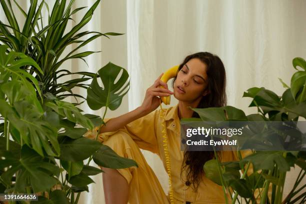 woman in yellow jumpsuit talking on yellow retro phone, sitting amount large monstera plants. - jumpsuit fashion stock pictures, royalty-free photos & images