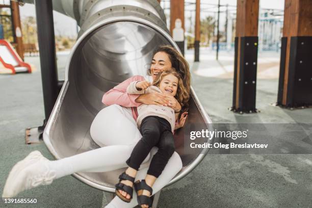 young happy mother with her daughter having fun in tube slide on playground. happy childhood and motherhood - child slide stockfoto's en -beelden