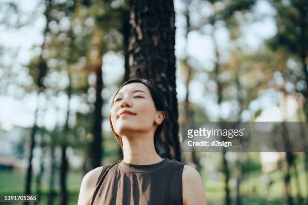 young asian woman with eyes closed leaning against tree in forest, with trees and sunbeam on background. enjoying fresh air and connection with nature. freedom in nature - ausdrucksstark stock-fotos und bilder