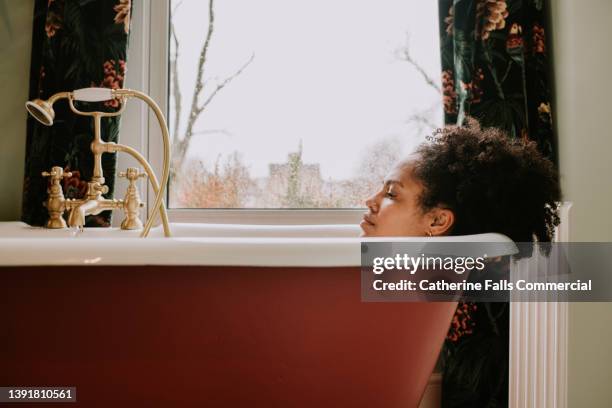 a beautiful woman bathes in a luxurious roll-top tub in front of a window, on a rainy day. - bathtub stock pictures, royalty-free photos & images