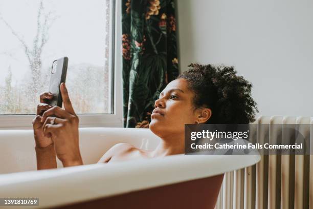 a relaxed woman uses her smart phone as she bathes in a luxurious roll-top bath - amenities hotel stockfoto's en -beelden