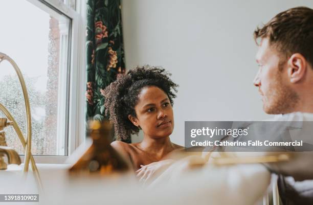 a couple have a conversation as the woman bathes and her partner leans on the side of the tub - couple bathtub photos et images de collection