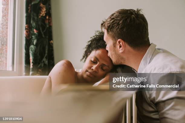 loving image of a couple in a bathroom. the woman lies in a bathtub and places her head gently on her partners shoulder as he kisses her forehead. - couple and kiss and bathroom 個照片及圖片檔