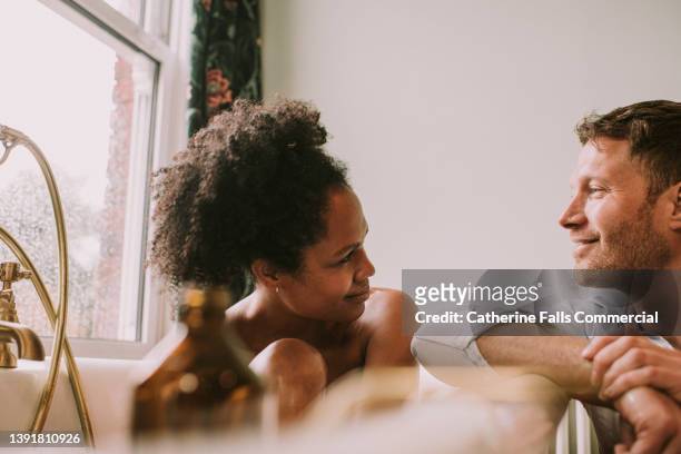a woman takes a hot bath as her partner rests his arm on the side of the tub and gazes at her lovingly. - couple and kiss and bathroom stock-fotos und bilder