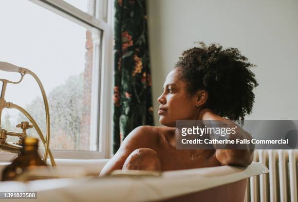 a beautiful woman daydreams in a roll top bathtub. she seems distracted as she gazes out the window. - distrait photos et images de collection