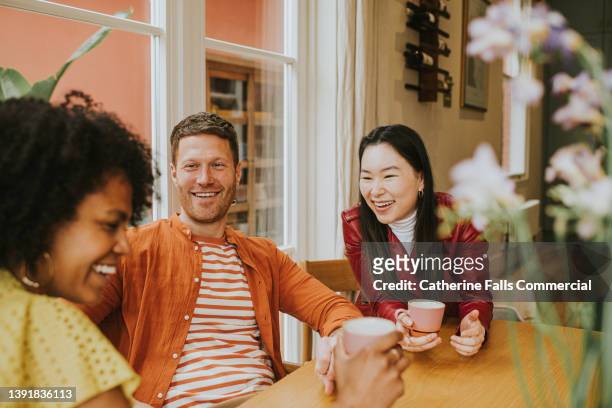 three young people sit around a table and giggle as they have a lighthearted discussion and drink coffee / tea - friendship stockfoto's en -beelden