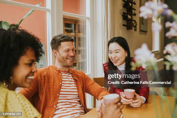 three young people sit around a table and giggle as they have a lighthearted discussion and drink coffee / tea - asian couple having hi tea stock pictures, royalty-free photos & images