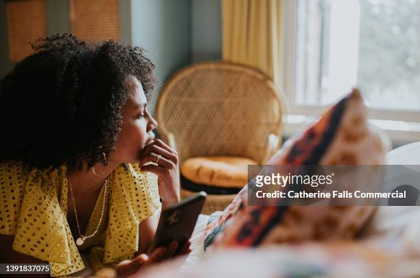 a woman looks distracted and pensive as she lies on a bed holding a mobile phone, and gazes out the window - beschouwing stockfoto's en -beelden