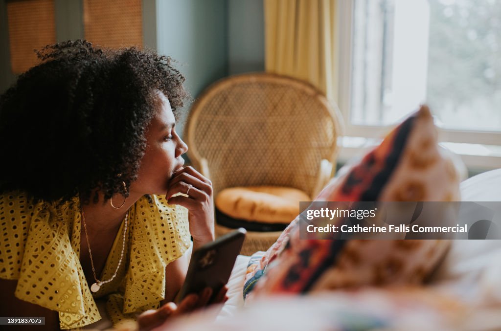 A woman looks distracted and pensive as she lies on a bed holding a mobile phone, and gazes out the window