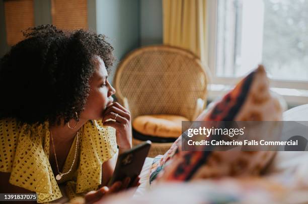 a woman looks distracted and pensive as she lies on a bed holding a mobile phone, and gazes out the window - mental burnout - fotografias e filmes do acervo