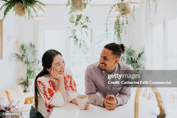 a woman and a man lean on a white surface in a bright, airy room, surrounded by houseplants. they smile at each other. - mental wellbeing stock pictures, royalty-free photos & images