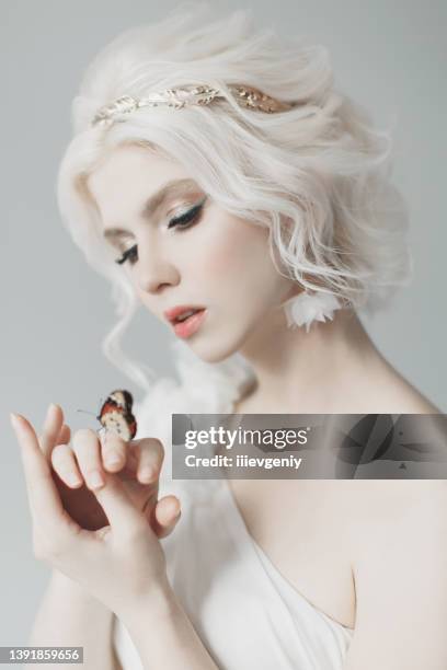 jeune mannequin blonde en studio. femme avec une belle coiffure grecque luxueuse en robe blanche. fille avec un papillon à la main. couronne d’or. albinos. portrait de beauté. déesse grecque. pureté et innocence - papillon fond blanc photos et images de collection