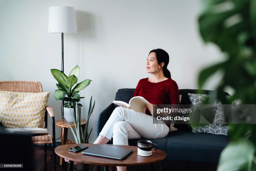 Carefree young Asian woman sitting on sofa reading a book and having coffee. Enjoying a quiet time and relaxing environment at cozy home