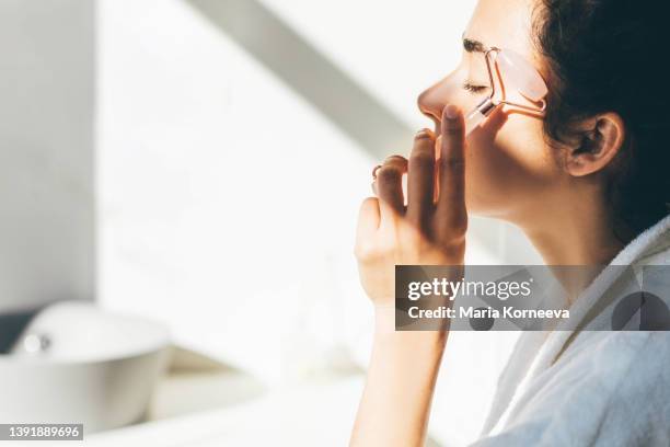 woman doing face massage with a roller at the bathroom. - facial massage stock pictures, royalty-free photos & images