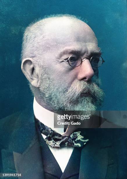 Colorized photograph of German scientist Robert Koch, from the chest up, wearing a dark suit, a bow tie, and wire-rimmed glasses, with his face...