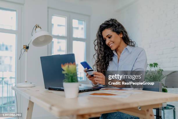 portrait of young woman holding credit card while sitting with laptop computer at the table at home - credit card debt stock pictures, royalty-free photos & images
