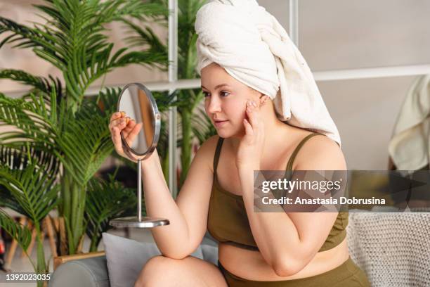 woman looking in mirror and touching face while siting on sofa - adult in mirror stock pictures, royalty-free photos & images