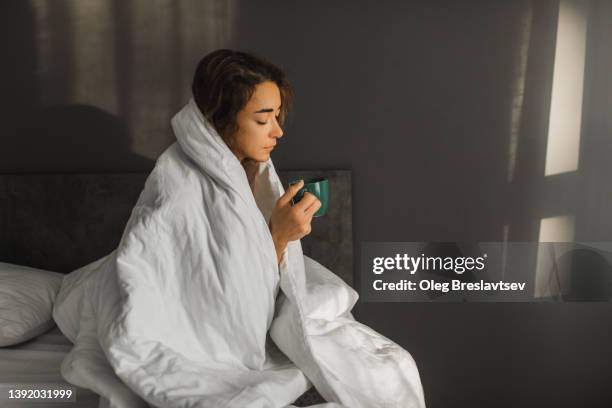 sad sick woman has a flu and drinking hot tea in bed and covered with blanket. cold morning, high temperature - hispanic person sick 個照片及圖片檔