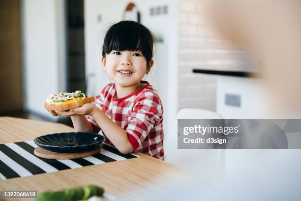 lovely smiling little asian girl sitting at dining table, chatting joyfully while having healthy breakfast with her mother in the morning. enjoying mother and daughter bonding time - mutter kind brot glücklich stock-fotos und bilder