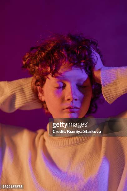 close-up portrait of a young woman with red short curly hair and freckles in a white sweater on a purple background, holding her head to with her eyes closed - touching face stock pictures, royalty-free photos & images