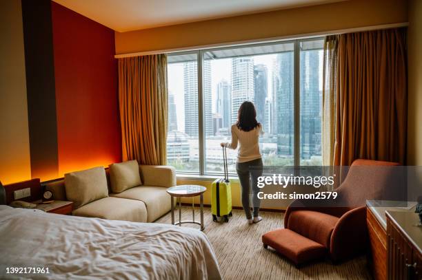 portrait of an asian tourist woman standing nearly window, looking to beautiful view with her luggage in hotel bedroom after check-in - hotel bedroom stock pictures, royalty-free photos & images