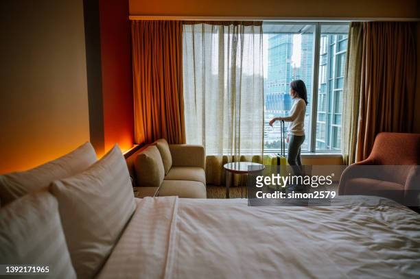 porträt einer asiatischen touristin, die fast am fenster steht und nach dem check-in mit ihrem gepäck im hotelzimmer eine schöne aussicht hat - gast stock-fotos und bilder