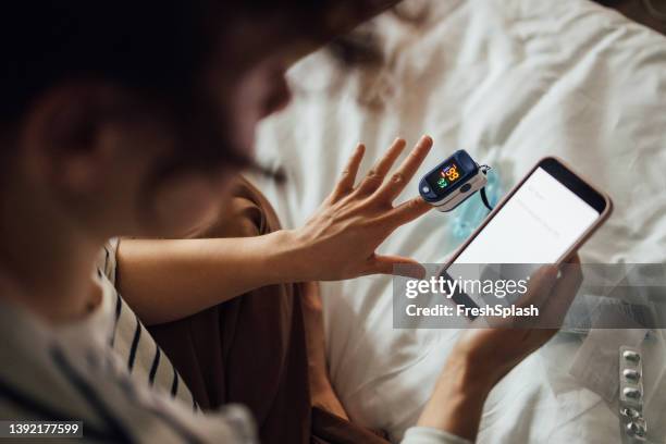 a from above shot of an unrecognizable woman measuring the oxygen saturation level of her blood after recovering from coronavirus while watching something on her smartphone - healthcare stock pictures, royalty-free photos & images