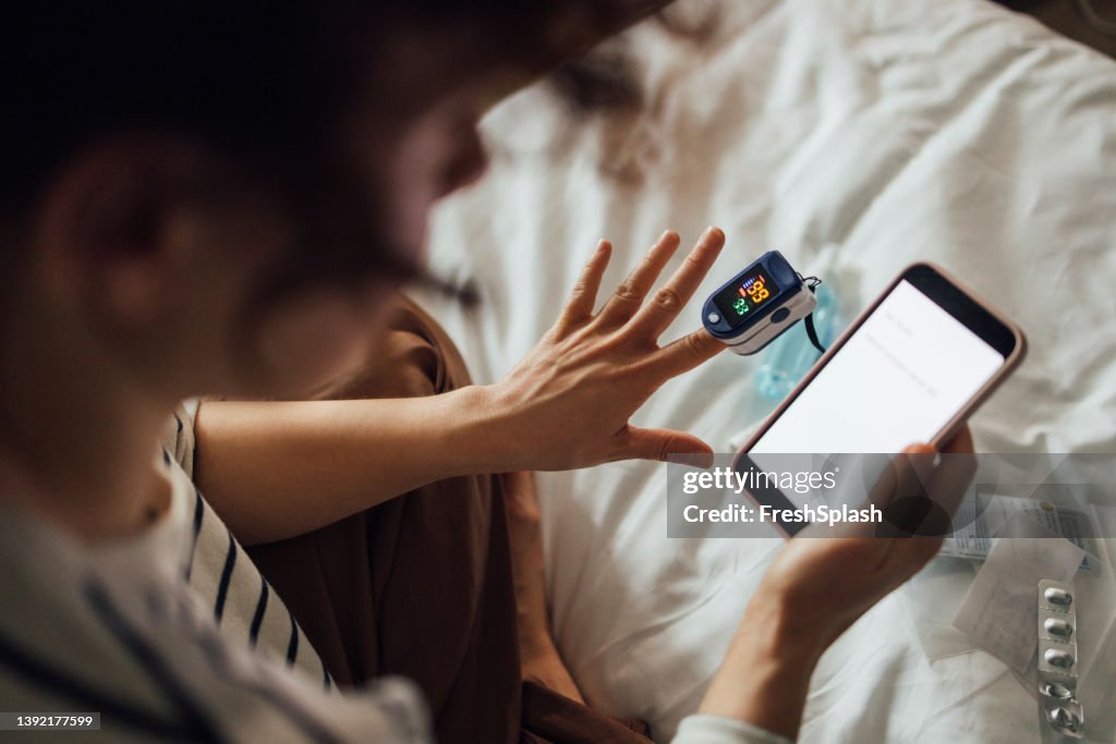 A From Above Shot Of An Unrecognizable Woman Measuring The Oxygen Saturation Level Of Her Blood After Recovering From Coronavirus While Watching Something On Her Smartphone