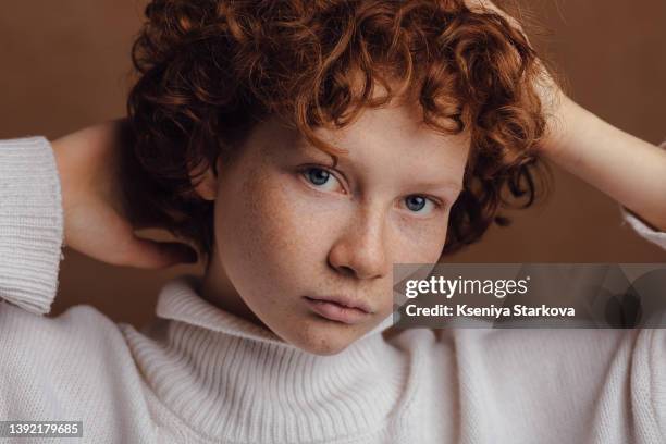 close-up portrait of a young woman with red short curly hair and freckles in a white sweater looks at the camera - sommersprossen stock-fotos und bilder