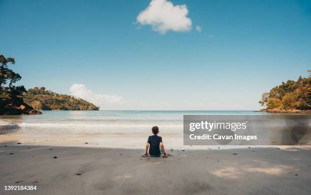 boy sitting alone on tropical beach looking at the ocean on sunny day. - costa rica beach stock pictures, royalty-free photos & images
