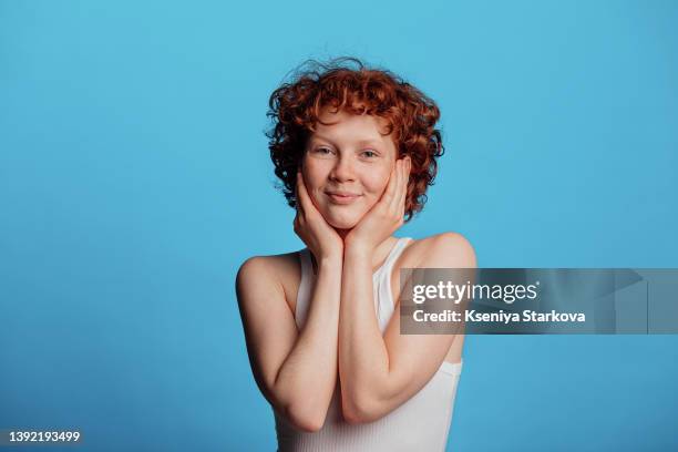 young woman with red short curly hair and freckles stands on a blue background, holds her face with her hands - girl with blue hair stock pictures, royalty-free photos & images