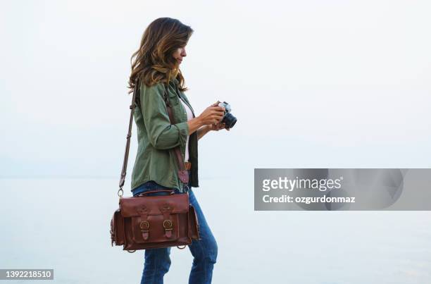 female photographer with a retro camera and leather bag near the beach - camera bag stock pictures, royalty-free photos & images