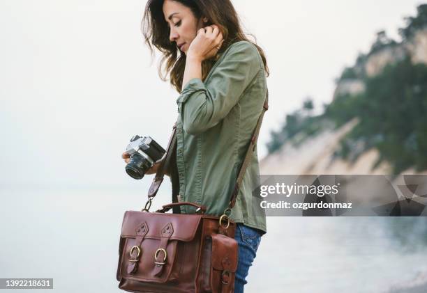 female photographer with a retro camera and leather bag near the beach - leather strap stock pictures, royalty-free photos & images