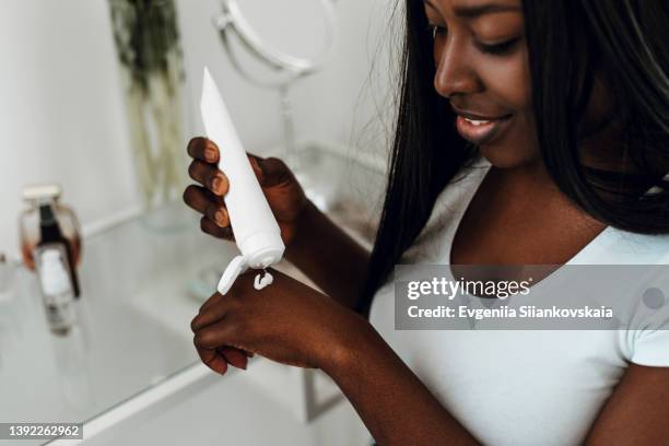 beautiful young african american woman holding cream tube and applying it at home. - creme tube imagens e fotografias de stock