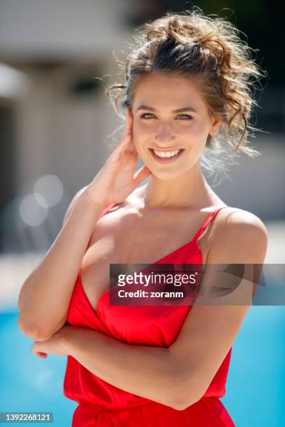 beautiful young fashion model wearing red sleeveless dress posing by the pool and smiling at camera - decolleté stockfoto's en -beelden